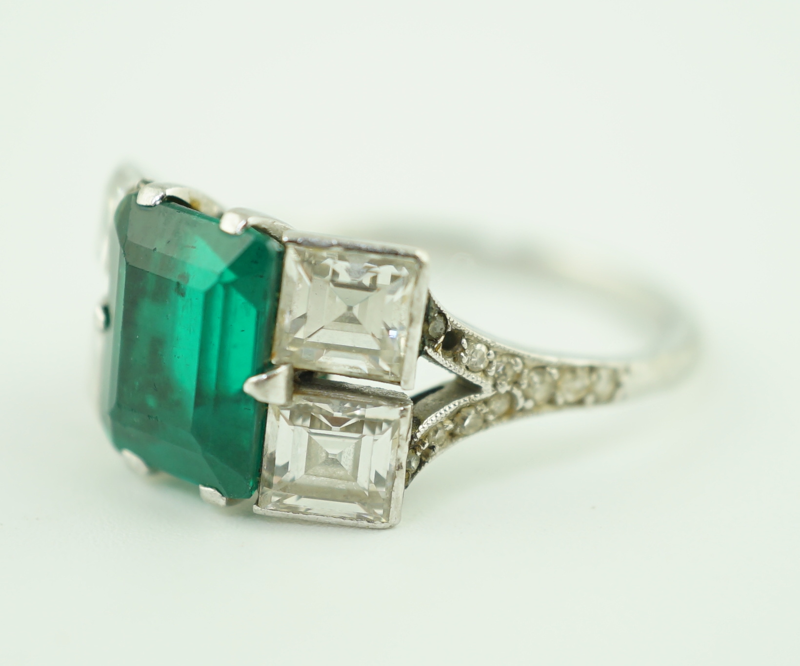 An Art Deco platinum, emerald and square cut diamond dress ring, with diamond chip set shoulders